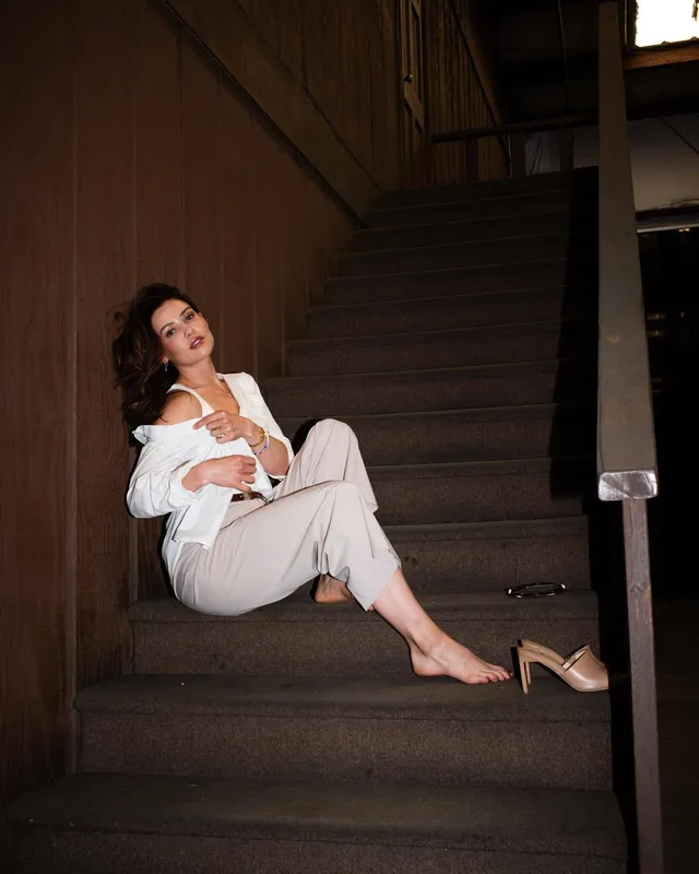 Danielle Campbell sitting on stairs in a white blouse and pale trousers.