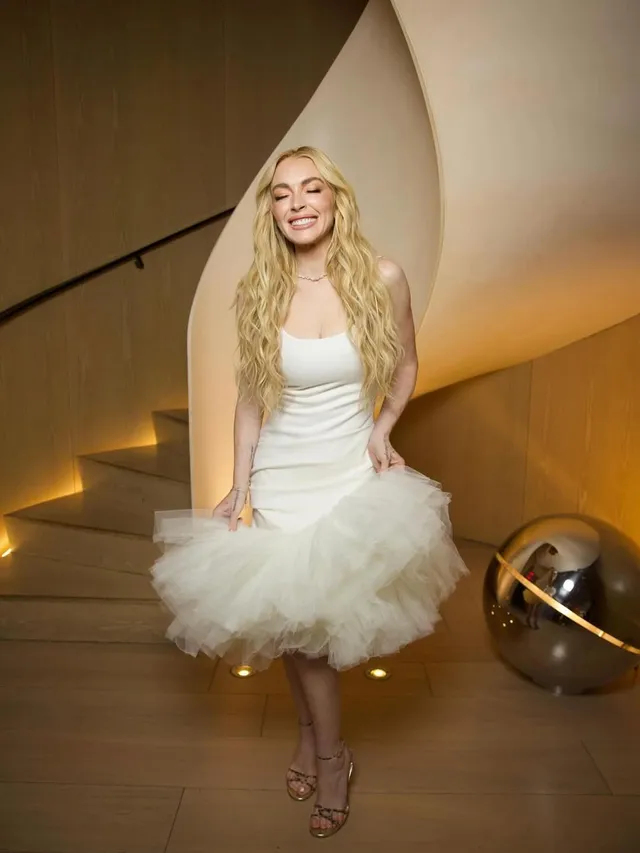 Lindsay Lohan posing indoors in a white dress and heeled sandals.