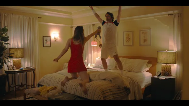 Kaley Cuoco jumping barefoot on a bed in a film scene.