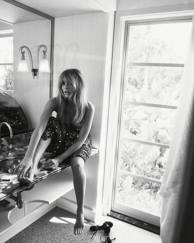 Margot Robbie sitting beside a bright window in a black-and-white fashion photograph.