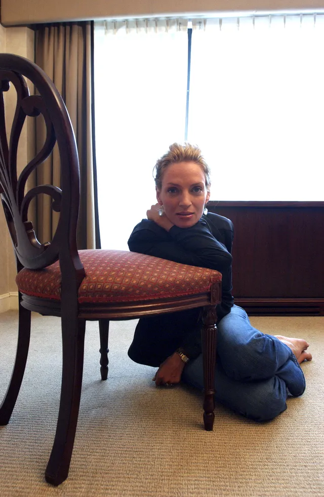 Uma Thurman sitting barefoot on the floor beside a chair.