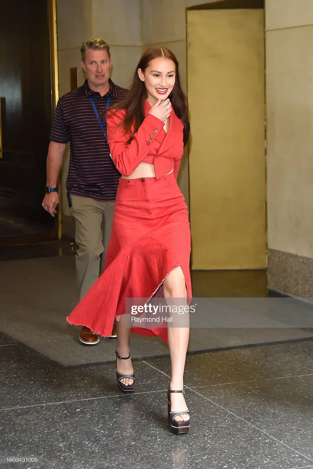 Lola Tung wearing a red skirt set and platform heels outside a building.
