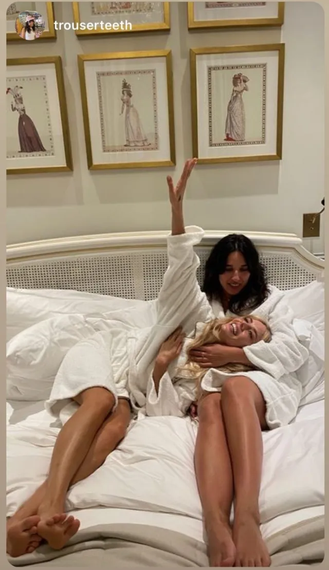 Samara Weaving reclining barefoot beside another woman on a bed.