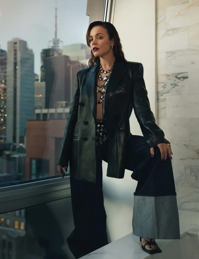 Rachel McAdams sitting beside a window in a suit and heels.