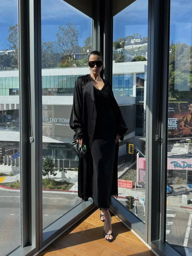 Adriana Lima standing in a dark outfit and heels beside a window.