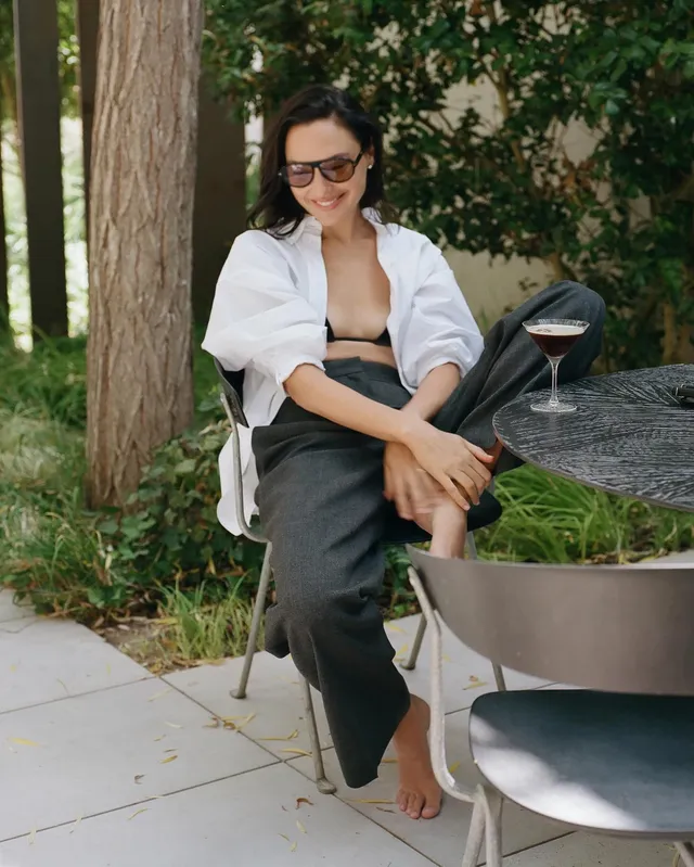 Gal Gadot sitting barefoot outside in a white shirt and dark trousers.