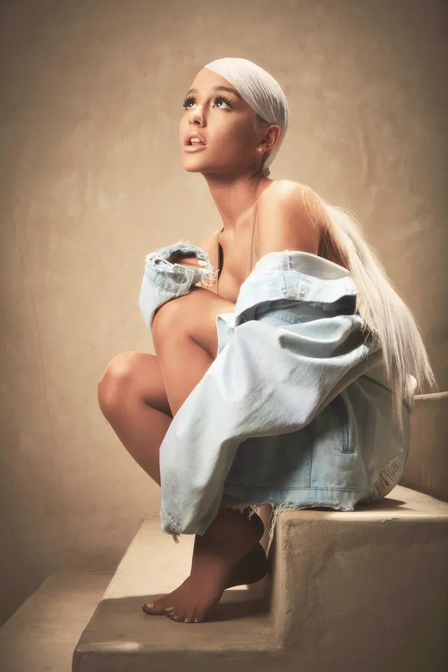 Ariana Grande: seated barefoot on a concrete step in a pale blue outfit.