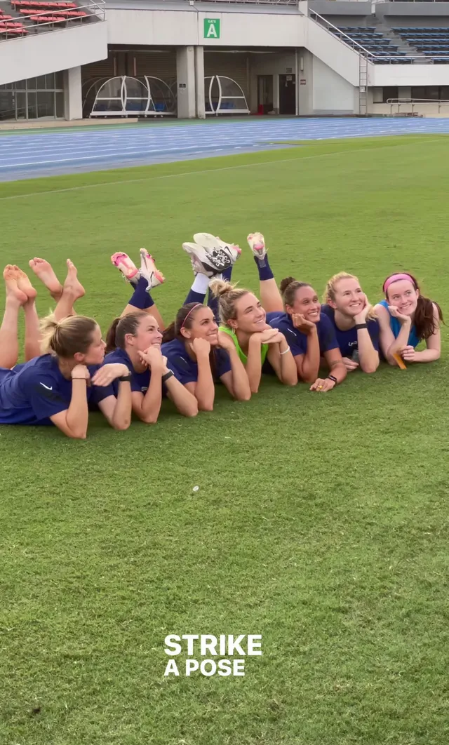 Alex Morgan posing on a sports field beside teammates with her bare feet raised.