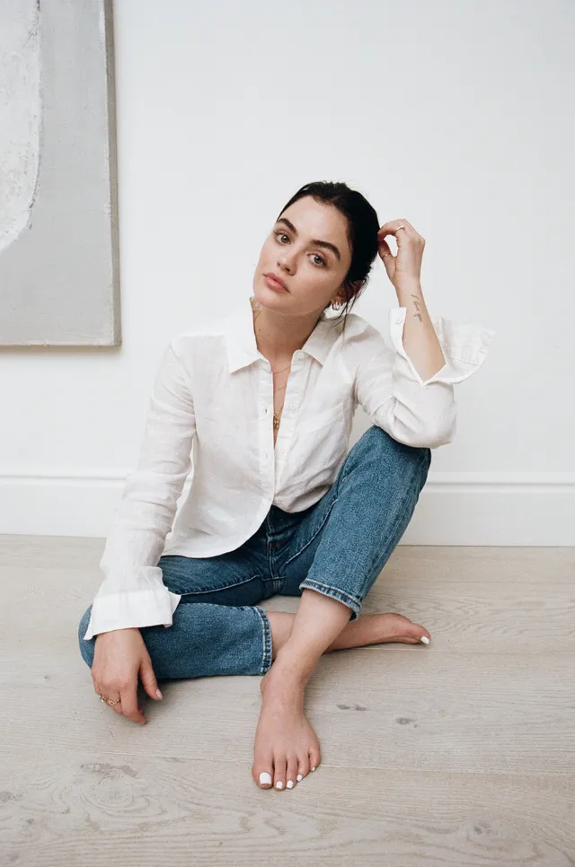 Lucy Hale sitting on the floor in a white blouse and jeans.