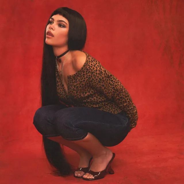 Ariana Greenblatt crouching in a patterned top and flat sandals against a red backdrop.