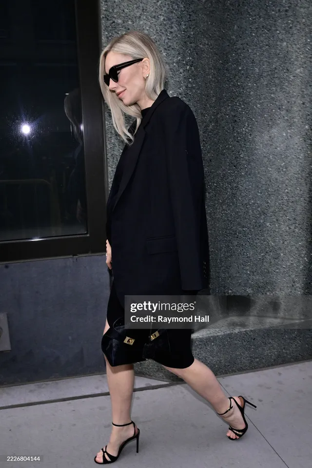 Vanessa Kirby walking beside a wall in a black dress and heels.