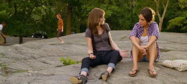 Mila Kunis sitting beside another adult in a patterned dress and heeled sandals.