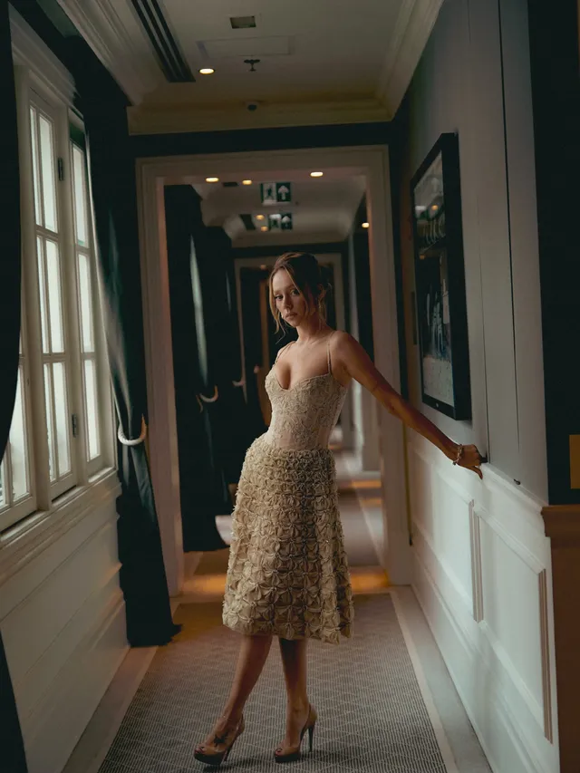Ester Expósito standing in black heels and a fitted patterned dress in a hallway.