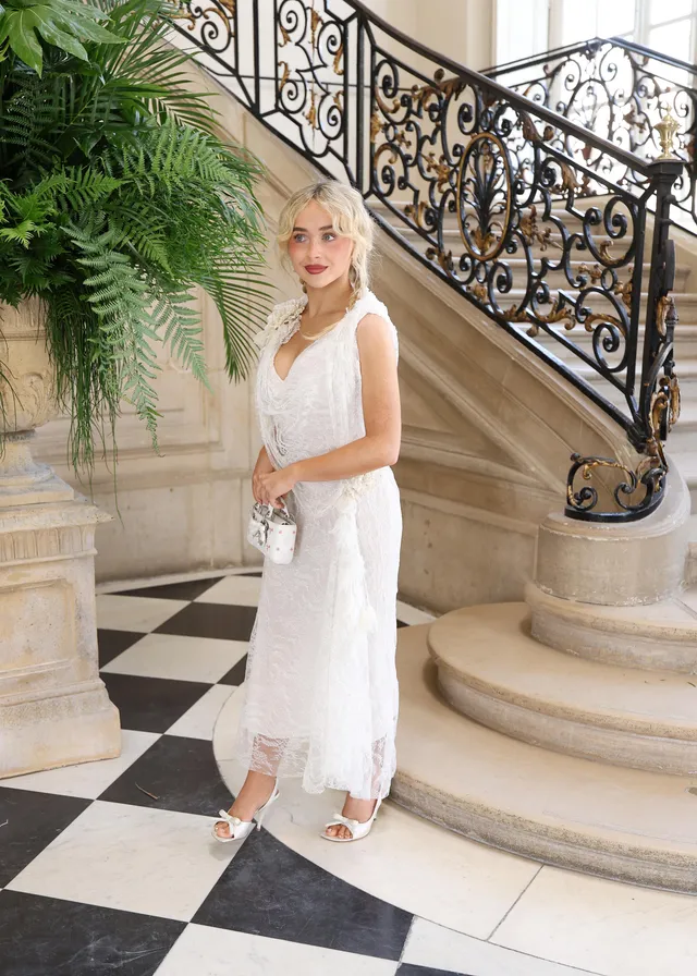 Sabrina Carpenter posing in a white lace dress and white heels indoors.