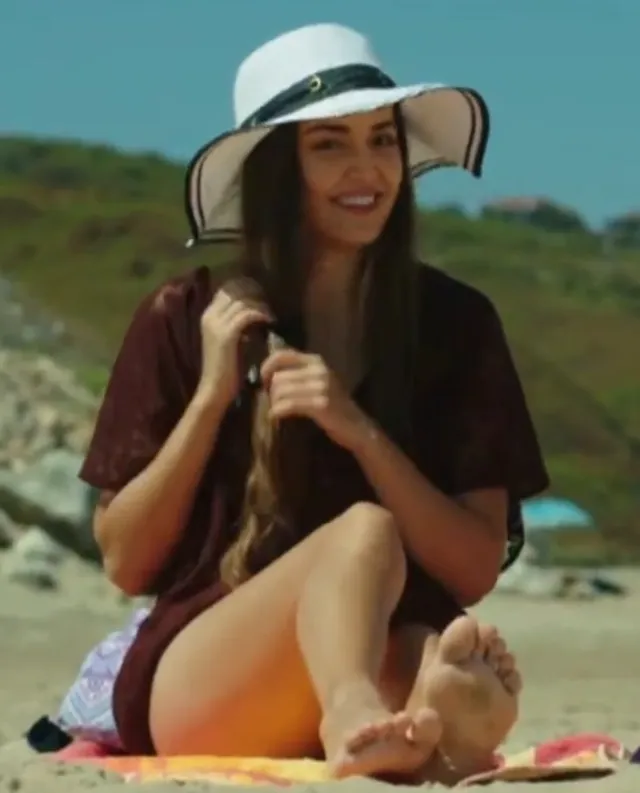 Hande Erçel sitting barefoot on a beach in a dark outfit and sun hat.