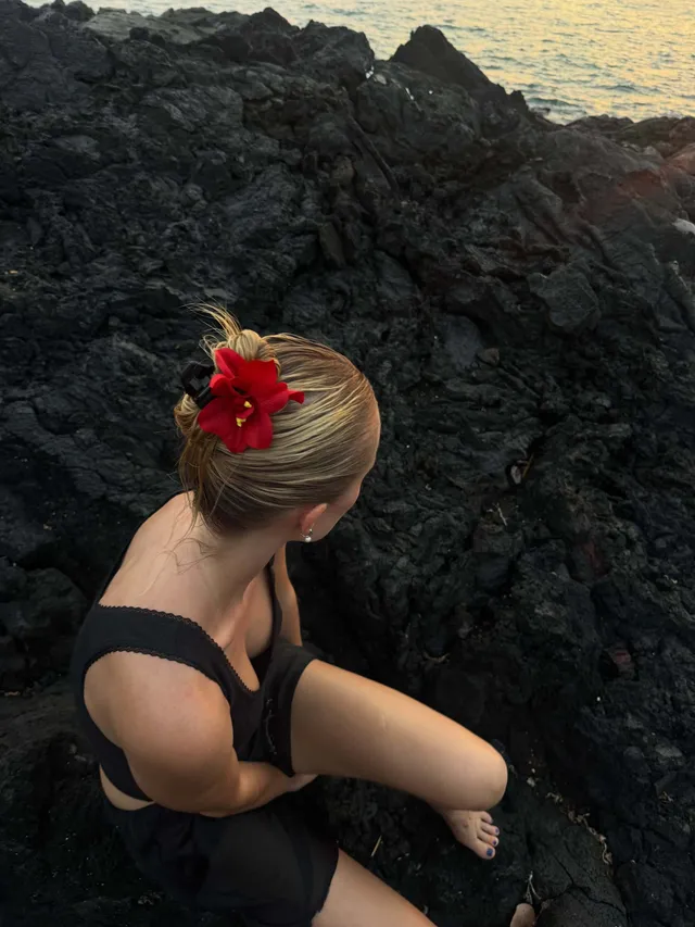 Maika Monroe sitting barefoot on coastal rocks.