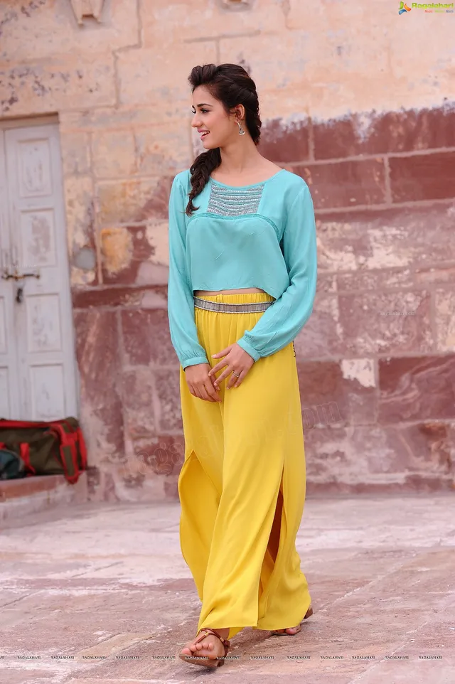 Disha Patani standing in flat sandals with yellow trousers and a turquoise blouse.