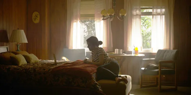 Anna Kendrick sitting beside a bed in a softly lit room.