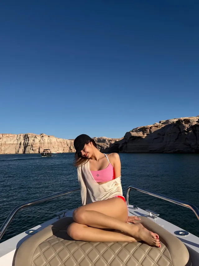 Lisa (Lalisa Manobal) sitting barefoot on a boat in a pale shirt and pink swimwear.