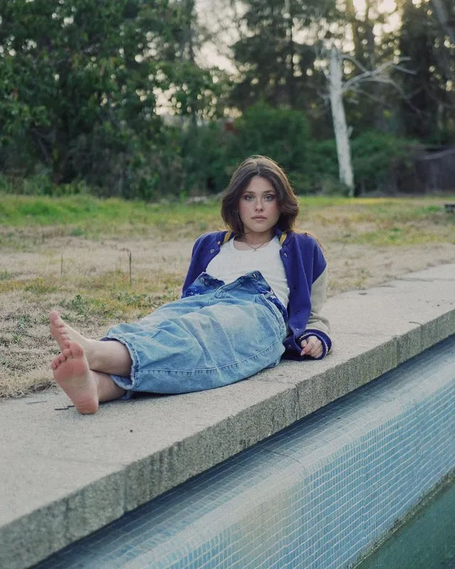 Nicole Wallace sitting barefoot beside a pool in jeans and a blue varsity jacket.