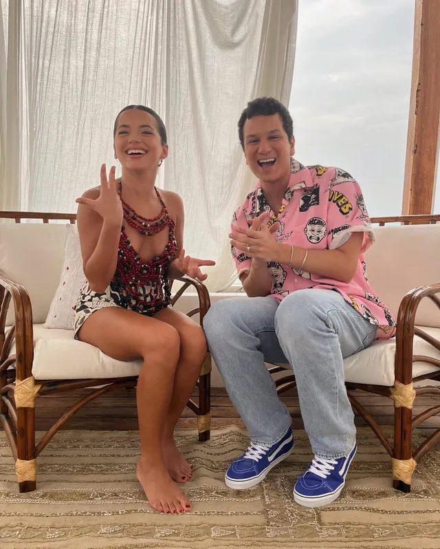 Isabela Merced sitting barefoot beside a man in a wicker chair.