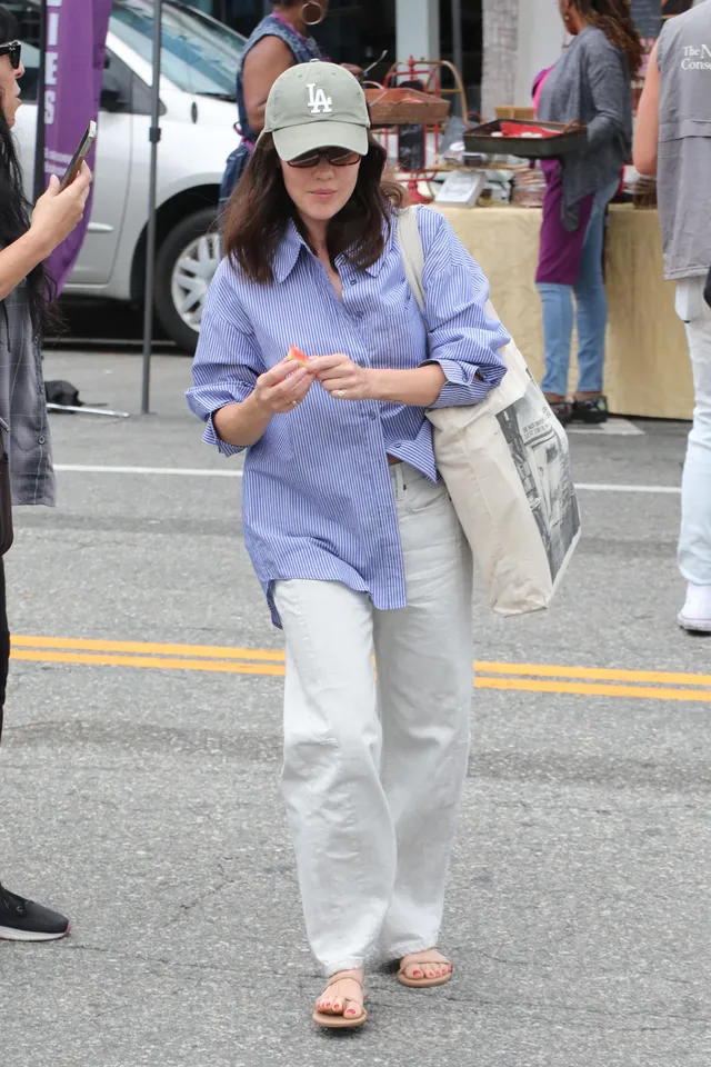 Lucy Hale walking beside a shop in light trousers and flip-flops.