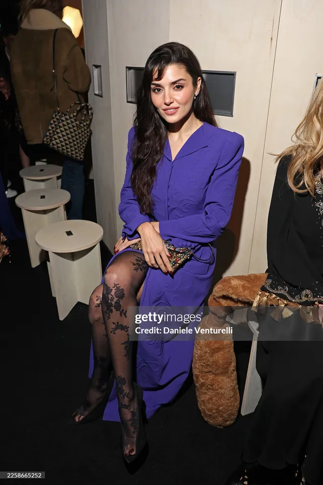 Hande Erçel sitting in a purple suit, patterned tights and black heels at an event.