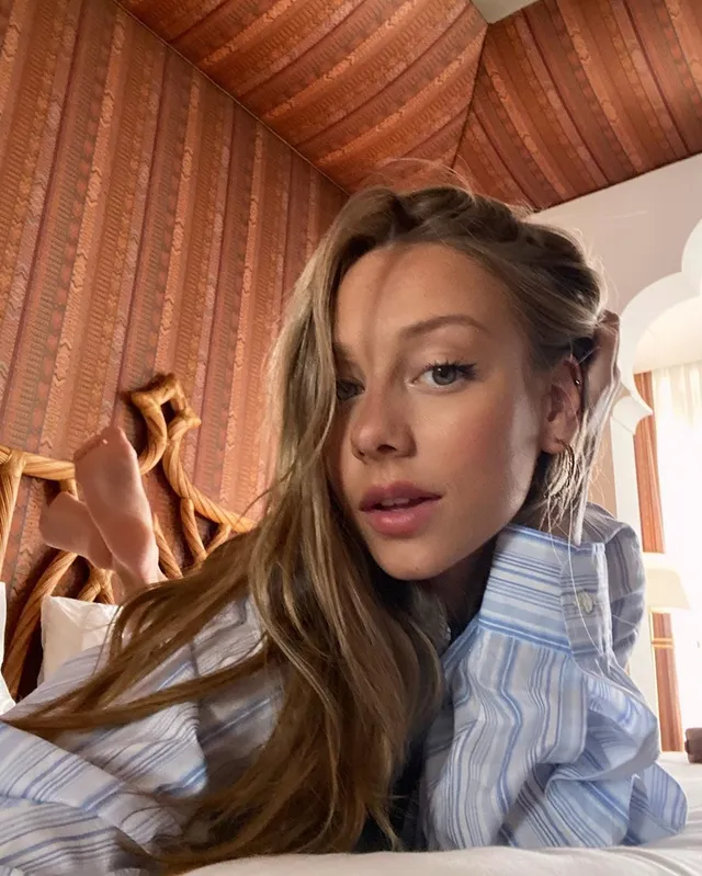 Ester Expósito reclining barefoot indoors in a striped shirt.