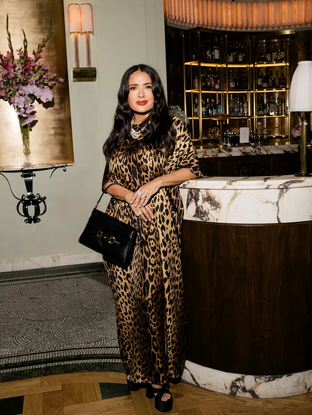 Salma Hayek standing in a leopard print dress beside a hotel counter.