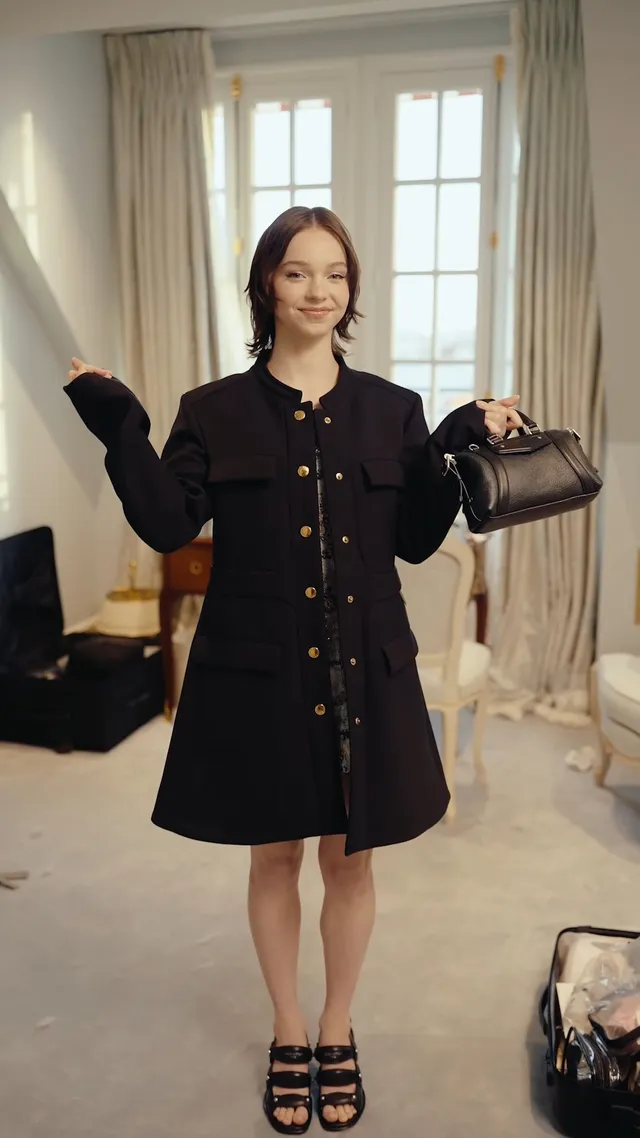 Emma Myers posing in a black dress and platform sandals indoors.