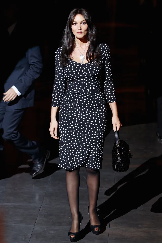 Monica Bellucci walking in a polka dot dress and black heels outdoors.