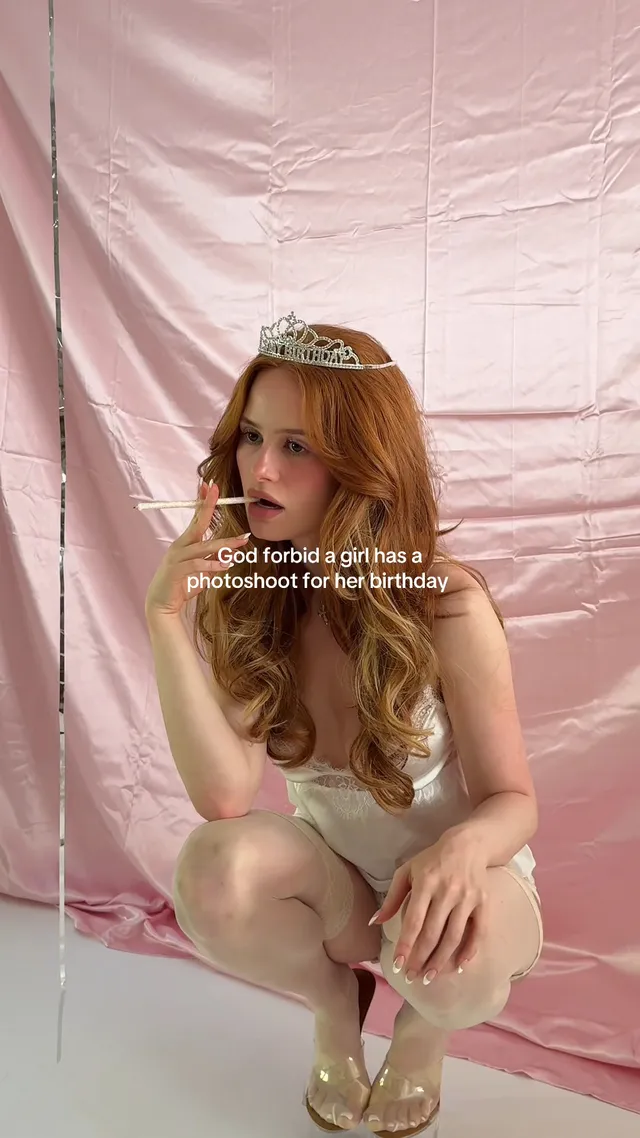 Madelaine Petsch crouching in pale socks and gold heels beside a pink backdrop.