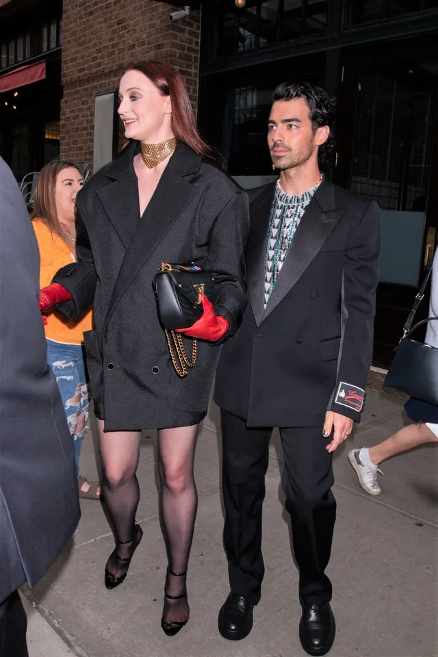 Sophie Turner walking beside a man wearing a black outfit and heeled shoes.