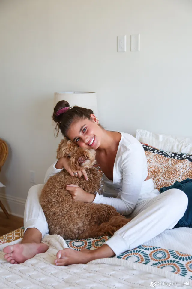 Taylor Hill sitting barefoot on a bed while holding a dog.