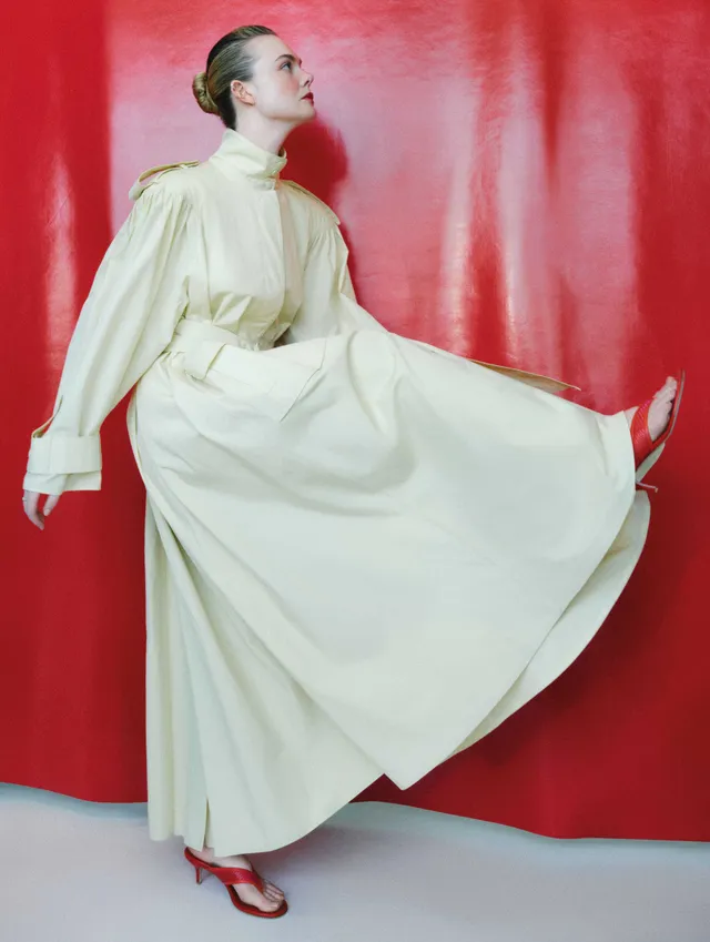 Elle Fanning posing against a red backdrop in a flowing white outfit and red heels.