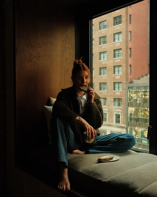 Sophie Nélisse sitting barefoot beside a window overlooking a city.
