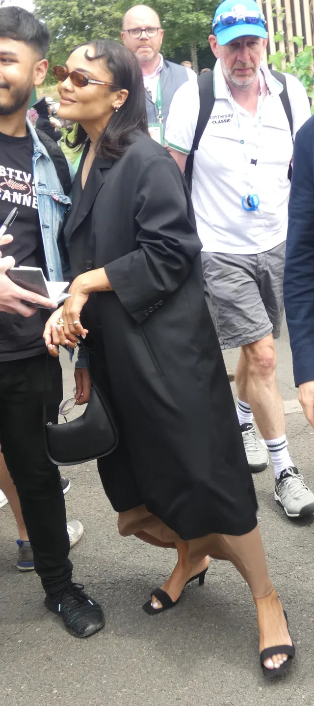 Tessa Thompson standing outdoors in a black coat and sandals.