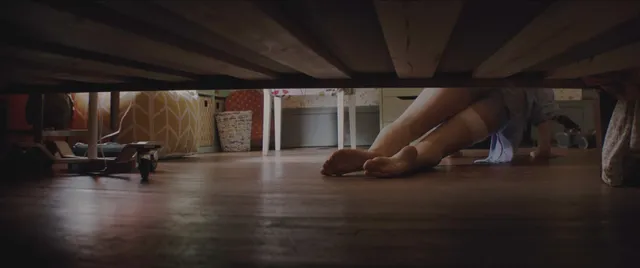 Chloë Grace Moretz with bare legs extended under a bed in a screen image.