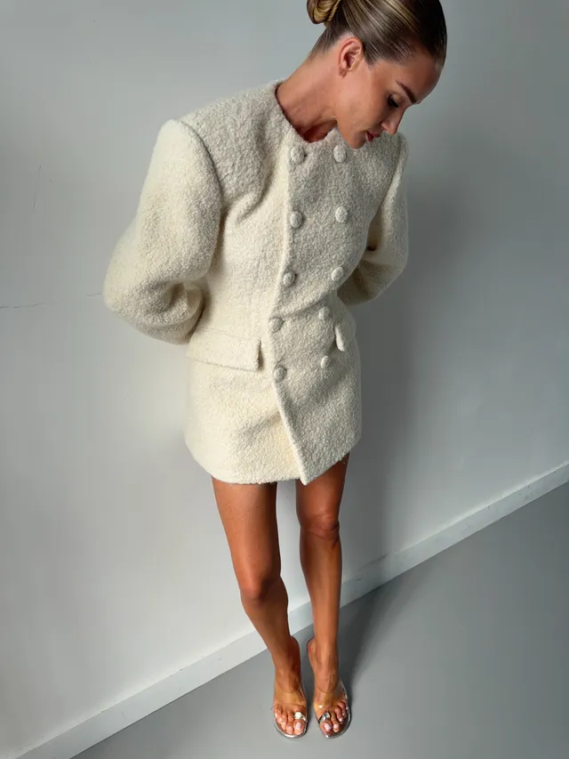Rosie Huntington-Whiteley standing in a pale jacket and gold heels indoors.
