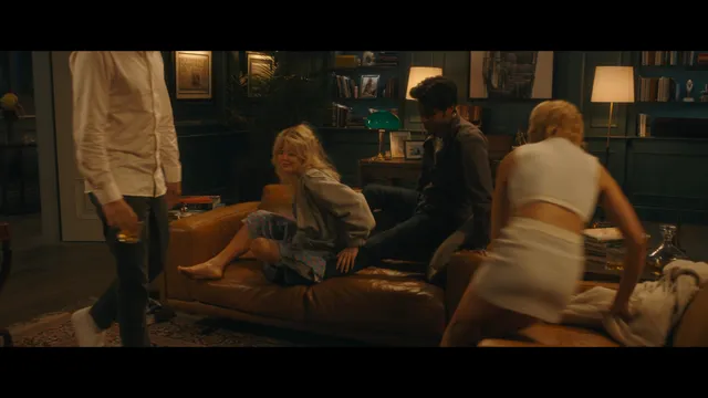 Emily Alyn Lind sitting on a sofa in a film scene with bare feet visible.