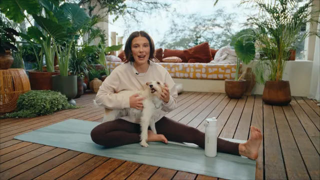 Millie Bobby Brown: seated barefoot on a yoga mat beside a small dog.