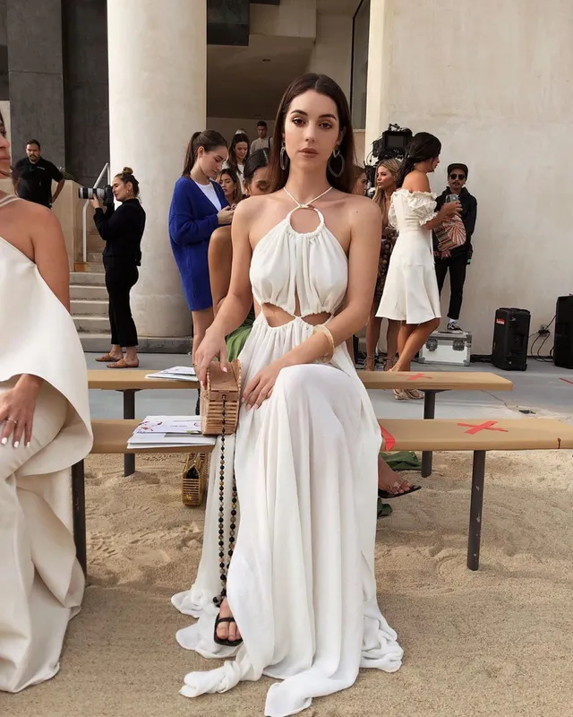 Adelaide Kane sitting at a fashion event in a long white dress.