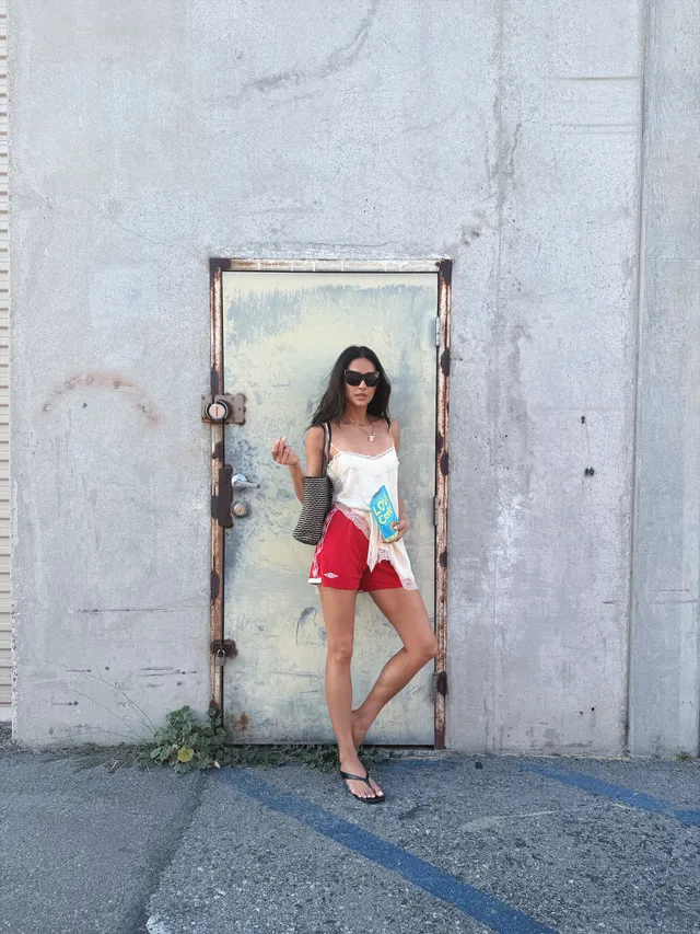 Shay Mitchell standing outside in a white top and red shorts.