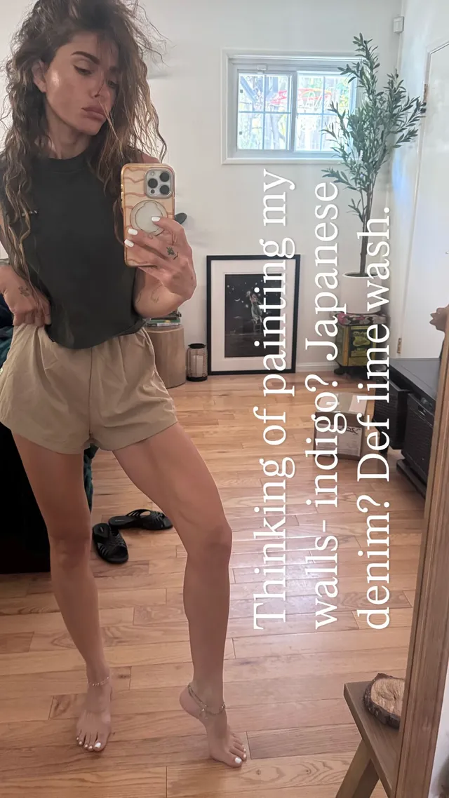 Sarah Shahi taking a mirror portrait in shorts and clear heels.