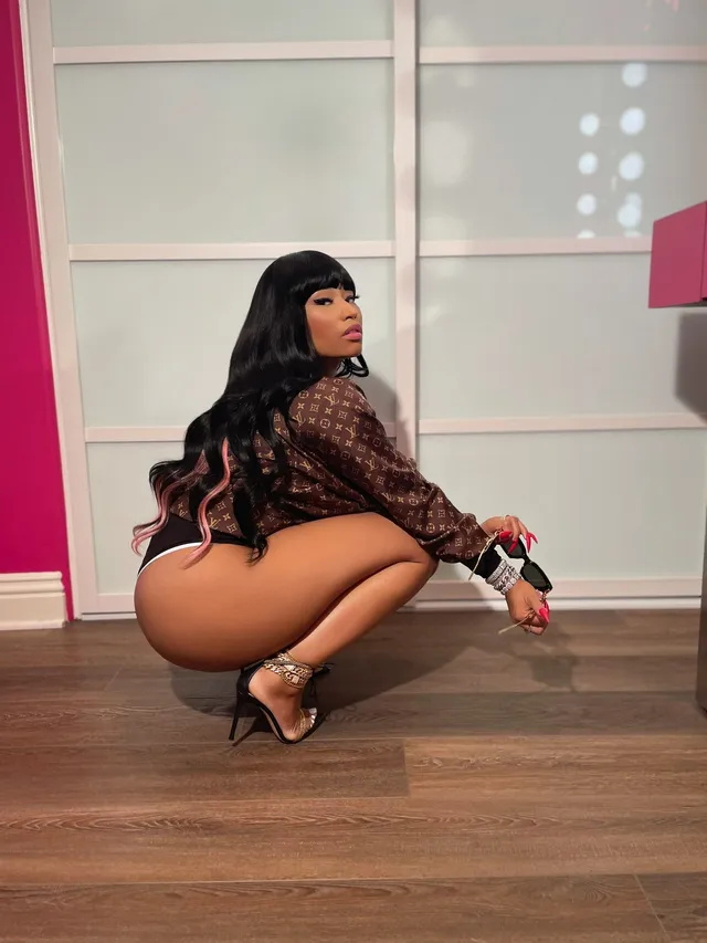 Nicki Minaj crouching indoors in a patterned top and black heels.