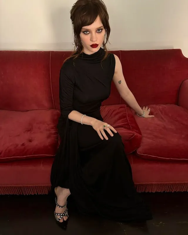 Sophie Thatcher sitting on a red sofa in a black dress and embellished heels.