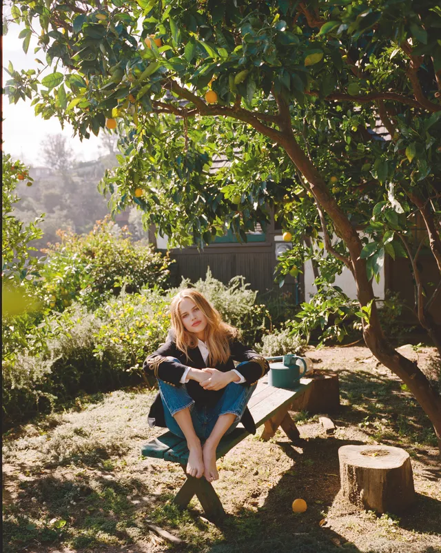 Halston Sage sitting barefoot outdoors on a bench beneath a tree.
