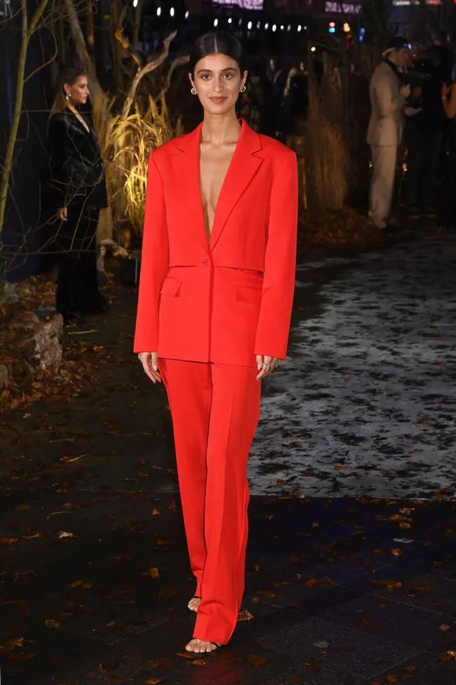 Anya Chalotra posing outdoors in a red trouser suit and heels.