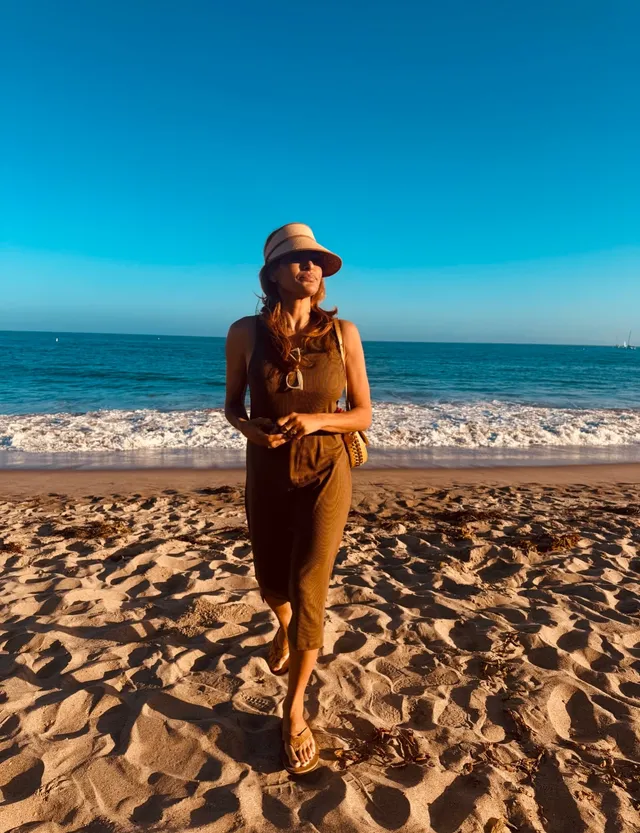 Eva Mendes standing on a beach in a brown outfit and flat sandals.