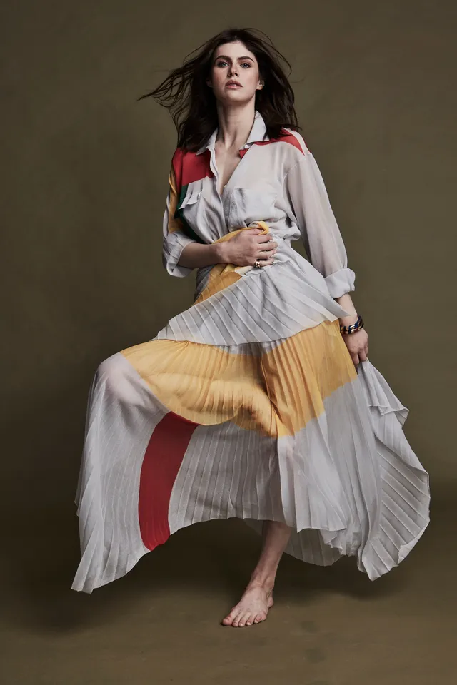 Alexandra Daddario posing barefoot in a flowing pleated dress.